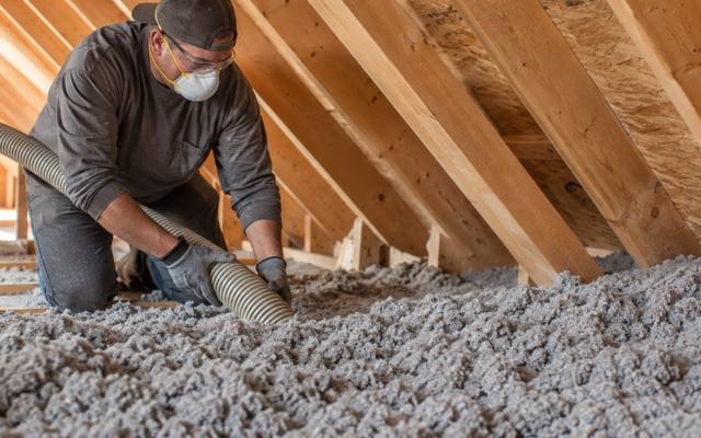 Worker holding a wide hose in grey loose-fill under the rafters while cleaning an attic