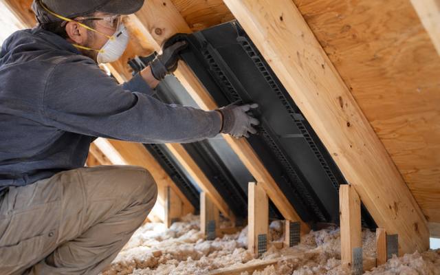 Worker fitting black ventilation baffles at the eaves before new attic insulation goes in